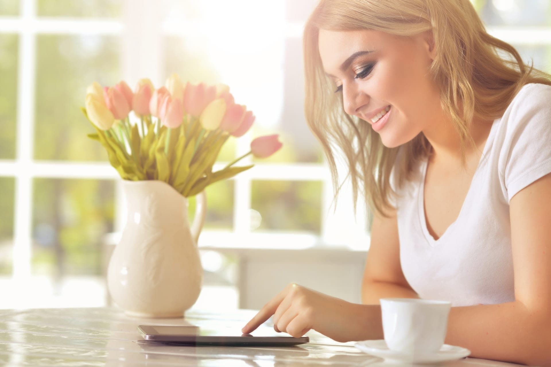 Lachende vrouw aan een zonnige tafel die op een tablet kijkt, met een vaas roze tulpen en een kop koffie ernaast